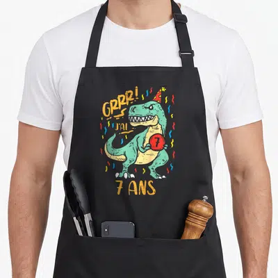 Tablier de cuisine noir avec illustration T-Rex vert portant une casquette et tenant un ballon 7 entouré de confettis multicolores