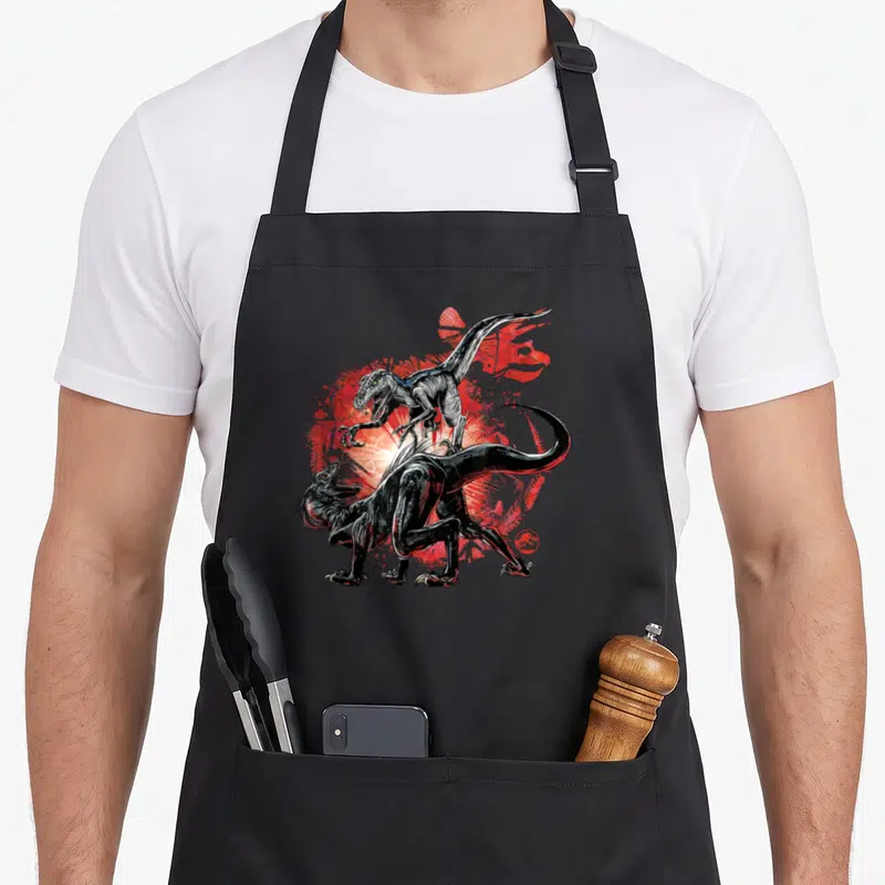 Tablier de cuisine noir avec illustration de combat entre deux dinosaures sur fond rouge éclaboussé.