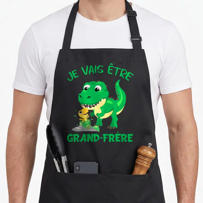 Tablier de cuisine noir avec un grand dinosaure vert et un petit dino jaune sortant d'une coque, texte vert en haut et en bas.