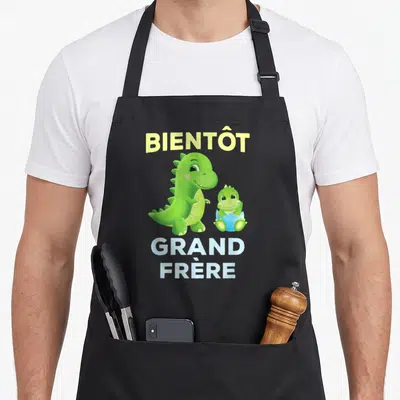 Tablier de cuisine noir avec deux dinosaures verts et le texte Bientôt Grand Frère imprimé en jaune et blanc.