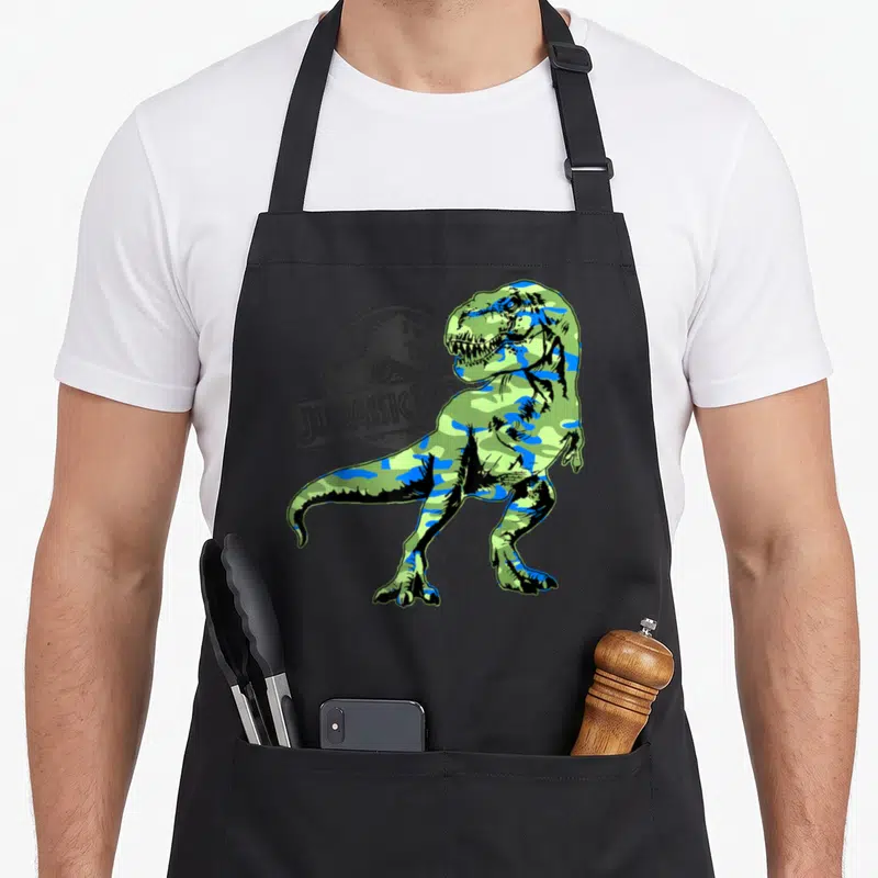 Tablier de cuisine noir avec un T-Rex vert et bleu en motif camouflage devant un logo circulaire gris.