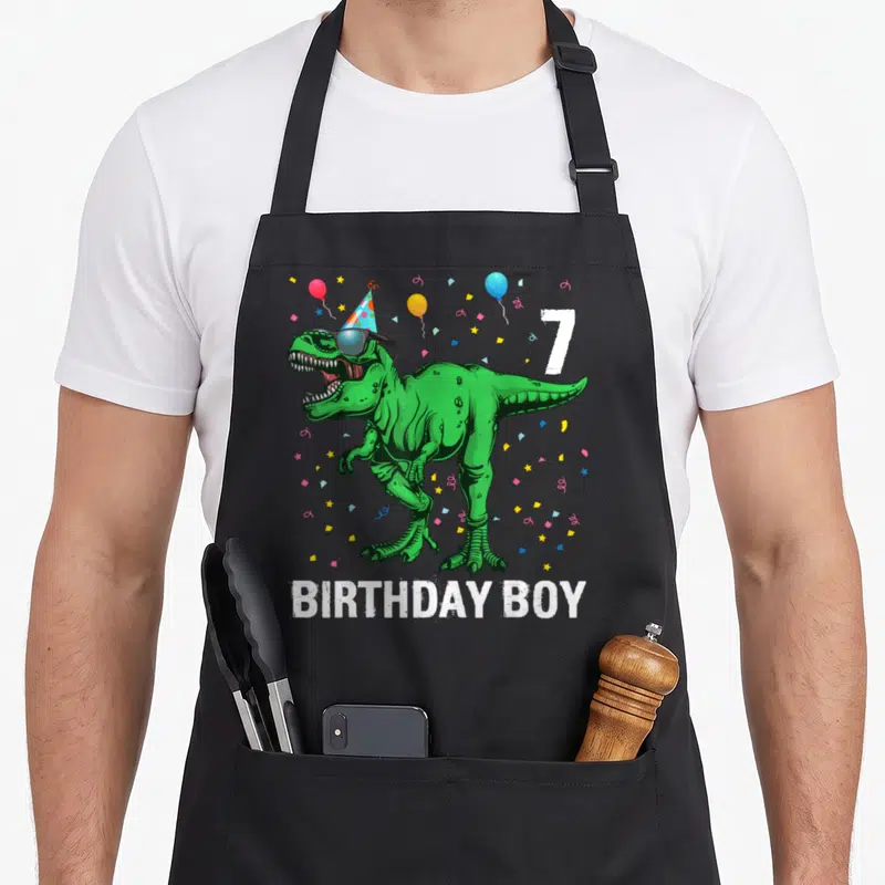 Tablier de cuisine noir avec un t-rex vert portant une casquette de fête et le texte anniversaire boy sept ans.