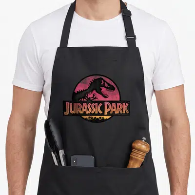Tablier de cuisine noir avec logo circulaire Jurassic Park et silhouette de dinosaure T-Rex sur fond rose.