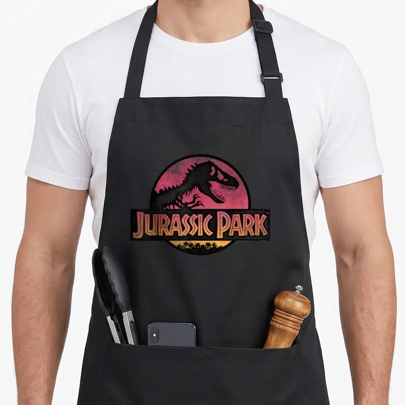 Tablier de cuisine noir avec logo circulaire Jurassic Park et silhouette de dinosaure T-Rex sur fond rose.