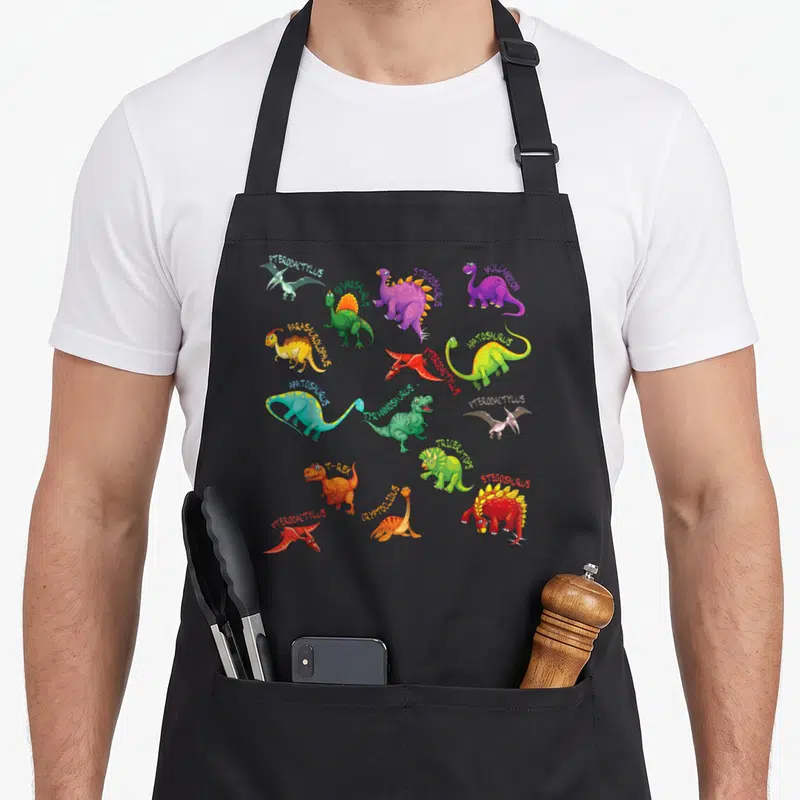 Tablier de cuisine noir avec plusieurs dinosaures colorés et leurs noms imprimés en typographie variée.