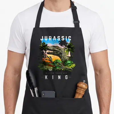 Tablier de cuisine noir avec illustration de dinosaure Jurassic King au milieu de palmiers.