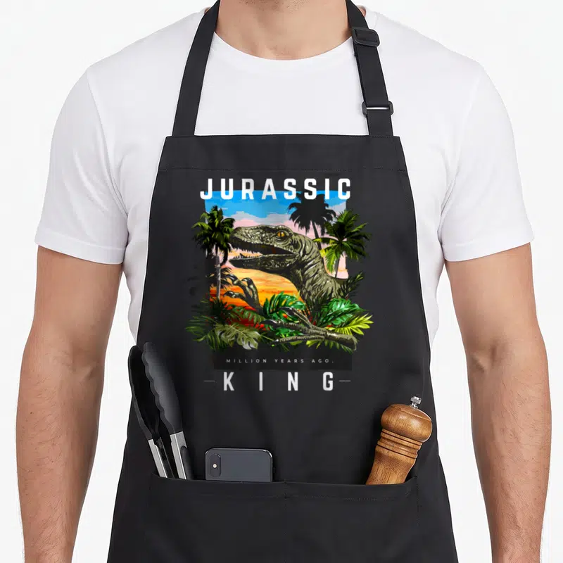 Tablier de cuisine noir avec illustration de dinosaure Jurassic King au milieu de palmiers.