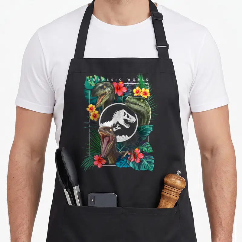 Tablier de cuisine noir avec trois têtes de raptors, fleurs roses et jaunes, feuilles vertes et logo T-Rex en cercle central.
