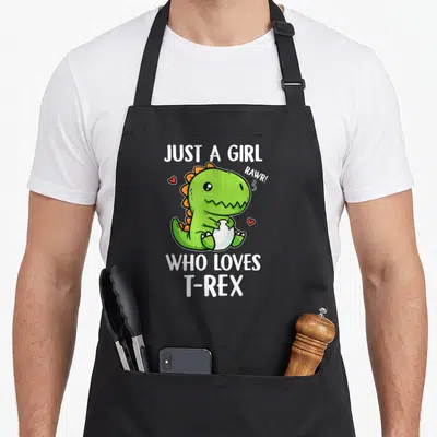 Tablier de cuisine noir avec un T-Rex vert tenant un œuf et le texte Just A Girl Who Loves T-Rex Rawr.