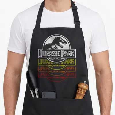 Tablier de cuisine noir avec logo Jurassic Park New Wold Fade en blanc, jaune et orange dégradé.