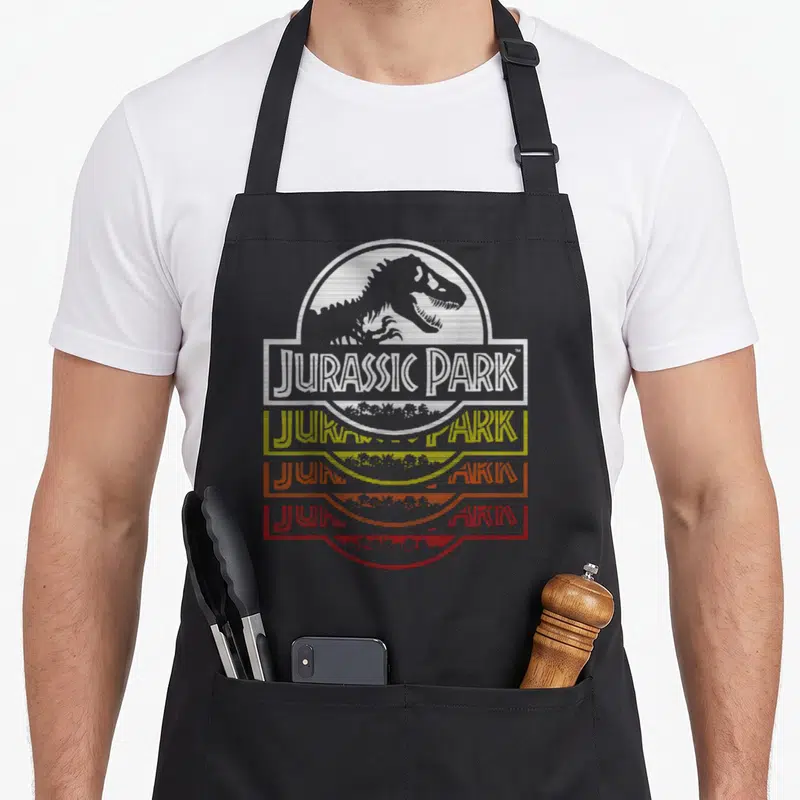 Tablier de cuisine noir avec logo Jurassic Park New Wold Fade en blanc, jaune et orange dégradé.