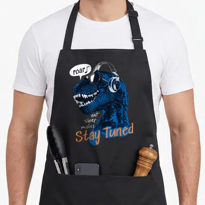 Tablier de cuisine noir avec illustration d'un T-Rex bleu portant des écouteurs et des lunettes de soleil, accompagné du texte Stay Tuned.