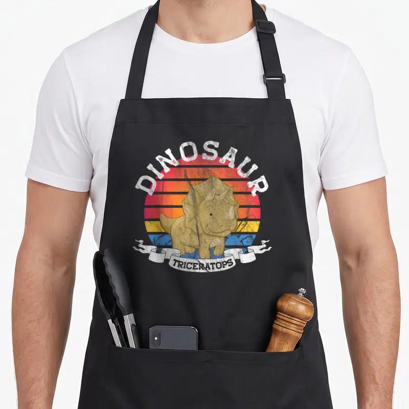 Tablier de cuisine noir avec illustration Triceratops souriant sur fond de soleil rétro coloré.