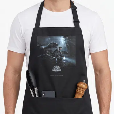 Tablier de cuisine noir avec illustration d'un homme et de dinosaures sous un projecteur lumineux, le logo Jurassic World en bas au centre.