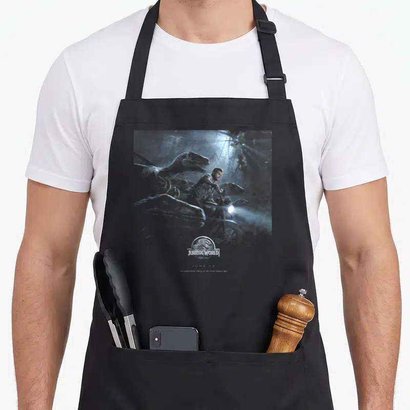 Tablier de cuisine noir avec illustration d'un homme et de dinosaures sous un projecteur lumineux, le logo Jurassic World en bas au centre.
