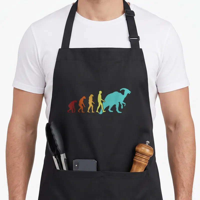 Tablier de cuisine noir avec une illustration colorée montrant l'évolution humaine menant à un dinosaure Parasaurolophus turquoise.