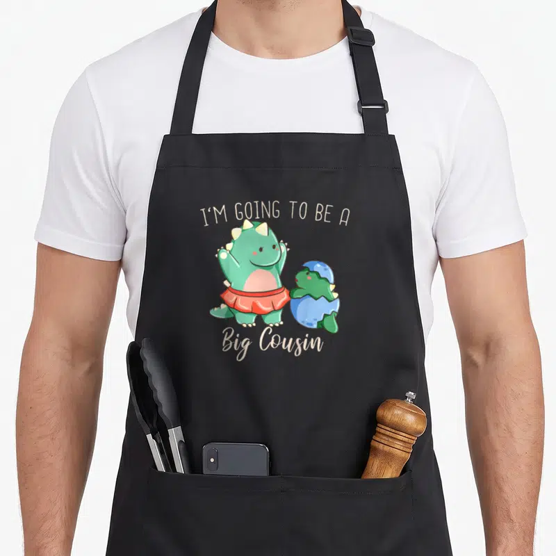 Tablier de cuisine noir avec un dinosaure vert en jupe rouge et un œuf bleu cassé.