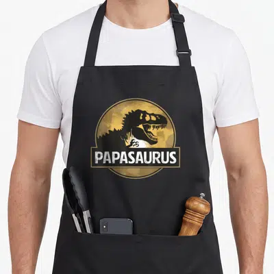 Tablier de cuisine noir avec logo circulaire doré affichant un T-Rex et le mot Papasaurus en blanc.