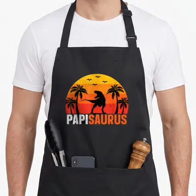 Tablier de cuisine noir avec illustration Papisaurus T Rex et palmiers sur fond dégradé orange.