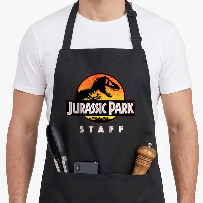 Tablier de cuisine noir avec logo circulaire orange et texte Jurassic Park STAFF en blanc.