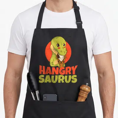 Tablier de cuisine noir avec un dinosaure vert tenant un sandwich et le texte HANGRY SAURUS en lettres colorées.