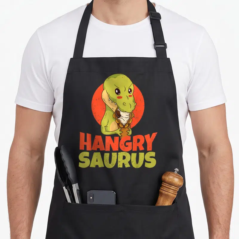 Tablier de cuisine noir avec un dinosaure vert tenant un sandwich et le texte HANGRY SAURUS en lettres colorées.