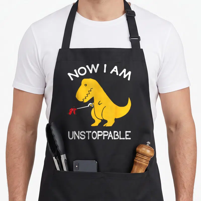 Tablier de cuisine noir avec un T-Rex jaune tenant une pince à crevettes et le texte Now I am unstoppable