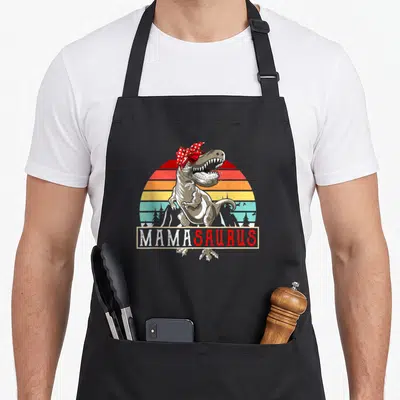 Tablier de cuisine noir avec illustration d'un T Rex portant un foulard à pois rouge et le texte MamaSaurus en bas.