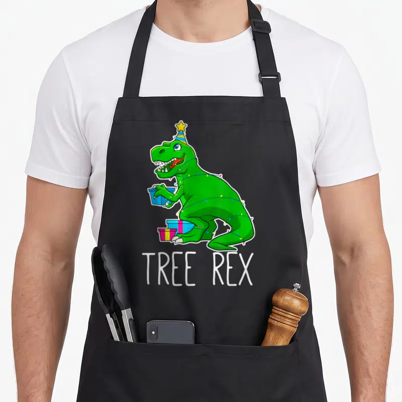 Tablier de cuisine noir avec un dinosaure vert en bonnet d'anniversaire tenant des cadeaux sous les guirlandes.