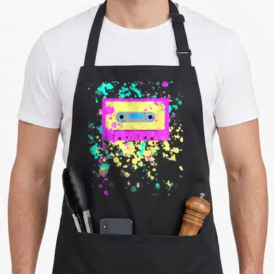 Tablier de cuisine noir avec illustration de cassette audio rose et jaune entourée d'éclaboussures colorées style graffiti.