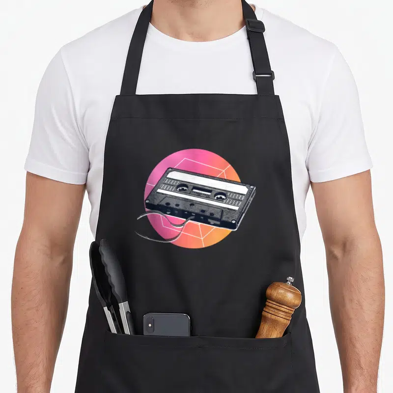 Tablier de cuisine noir avec illustration de cassette audio vintage sur fond géométrique rose et orange.