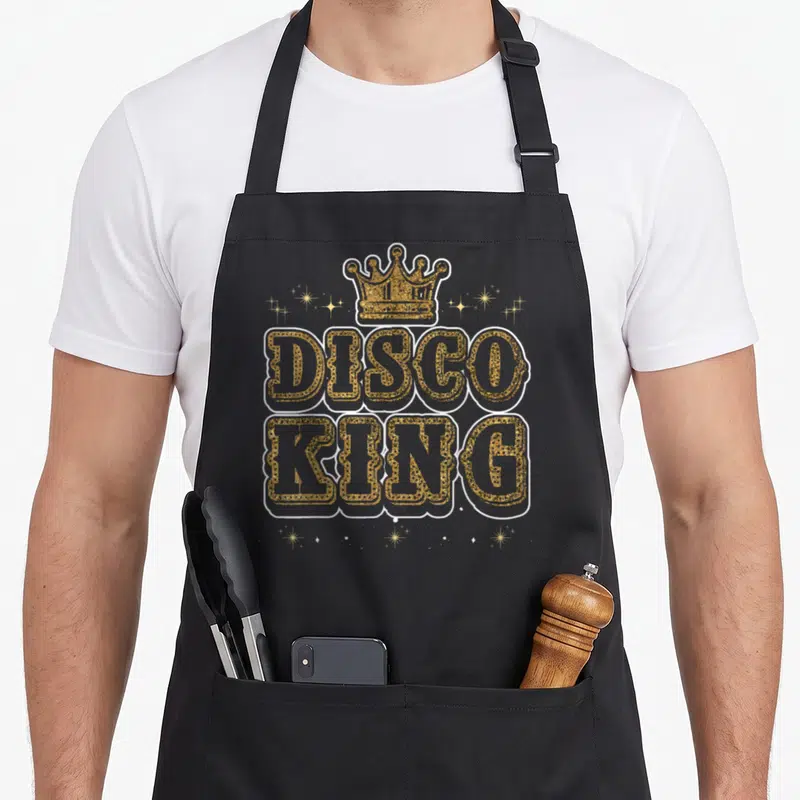 Tablier de cuisine noir avec motif Disco King en lettres dorées pailletées surmontées d'une couronne et d'étoiles scintillantes.