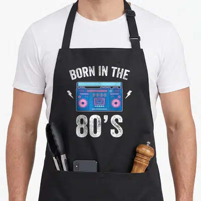 Tablier de cuisine noir avec une illustration de boombox bleue et rose et le texte Born in the 80's en blanc.