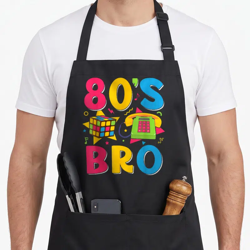 Tablier de cuisine noir avec typographie 80's Bro, téléphone vert et cube de Rubik coloré sur fond gris uni.