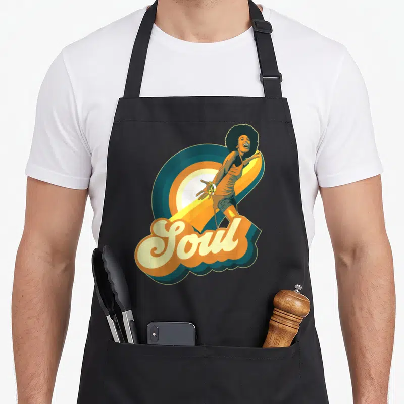 Tablier de cuisine noir avec une femme à l'afro chantant devant un disque vinyl et le mot Soul en lettres arrondies orange et bleu.