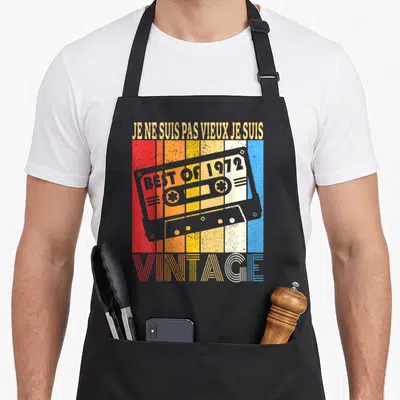 Tablier de cuisine noir avec illustration cassette audio vintage et texte humoristique sur fond rayé coloré.