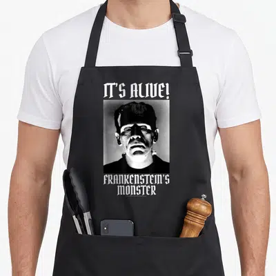 Tablier de cuisine noir avec portrait monochrome du monstre de Frankenstein et texte It's Alive en typographie gothique blanche.