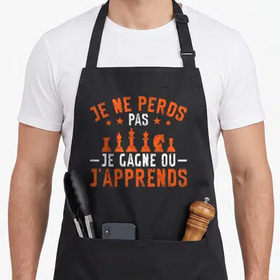 Tablier de cuisine noir avec inscription Je ne perds pas je gagne ou j'apprends et silhouettes de pièces d'échecs oranges.