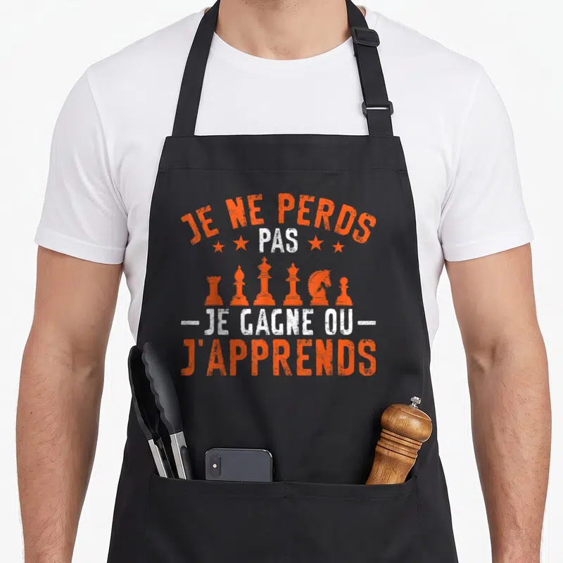 Tablier de cuisine noir avec inscription Je ne perds pas je gagne ou j'apprends et silhouettes de pièces d'échecs oranges.