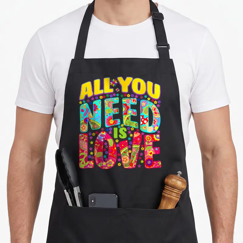 Tablier de cuisine noir avec inscription All You Need Is Love ornée de motifs floraux multicolores et d'ambiance flower power.
