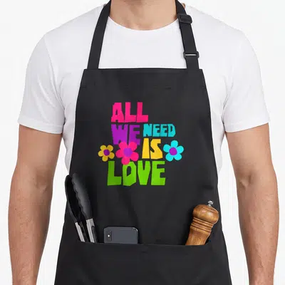 Tablier de cuisine noir avec l'inscription All We Need Is Love entourée de fleurs multicolores style années 70.