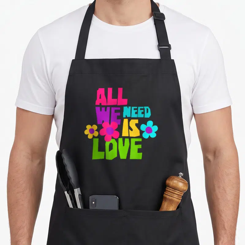 Tablier de cuisine noir avec l'inscription All We Need Is Love entourée de fleurs multicolores style années 70.