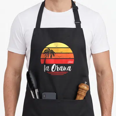 Tablier de cuisine noir avec un soleil tahitien rayé, une palme et le texte la Orana en blanc.