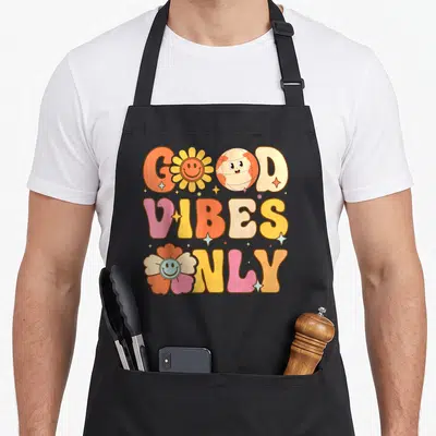 Tablier de cuisine noir avec inscription Good « Vibes » Only en lettres colorées, fleur et globe souriants style rétro.