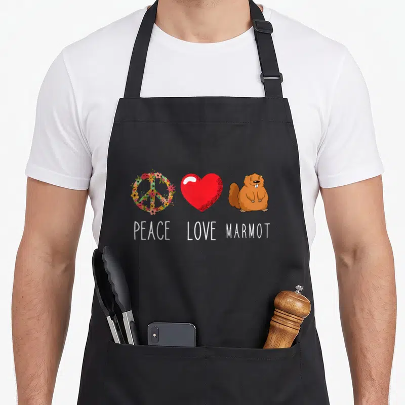 Tablier de cuisine noir avec un symbole de paix floral, un cœur rouge et une marmotte souriante sous les mots Peace Love Marmot.