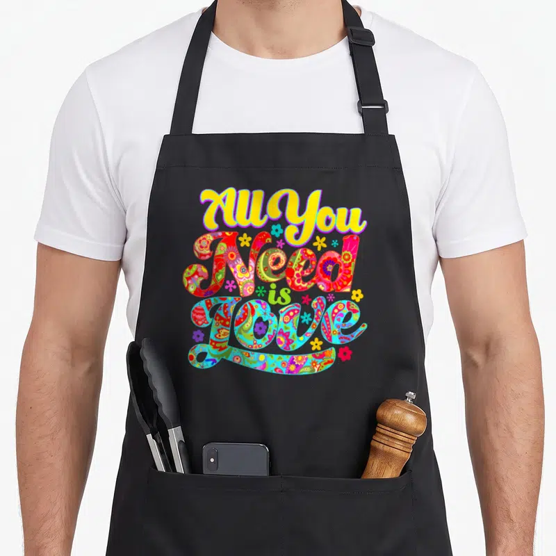 Tablier de cuisine noir avec l'inscription All You Need Is Love en lettres colorées et motifs floraux hippie.