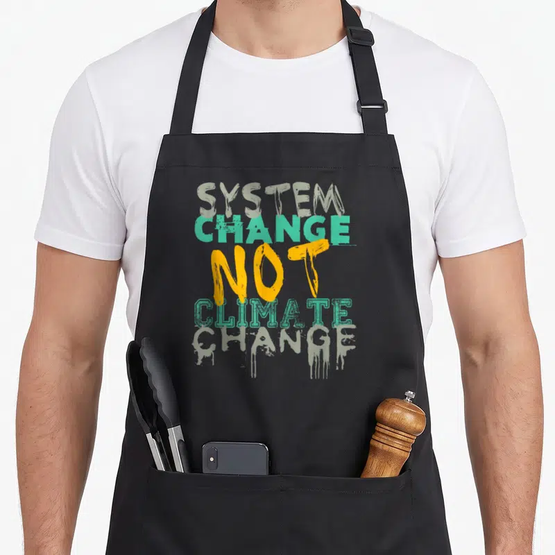 Tablier de cuisine noir avec inscription System Change Not Climate Change en lettres vertes et jaunes style graffiti.