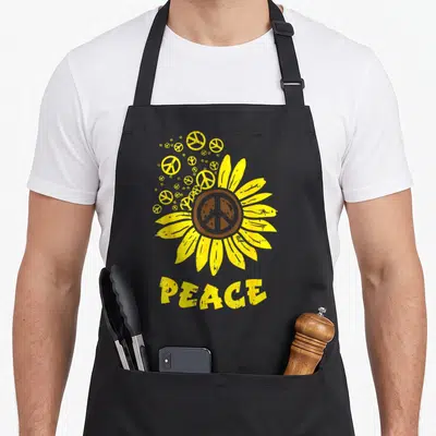 Tablier de cuisine noir avec un grand tournesol jaune stylisé et le mot PEACE imprimé en bas.