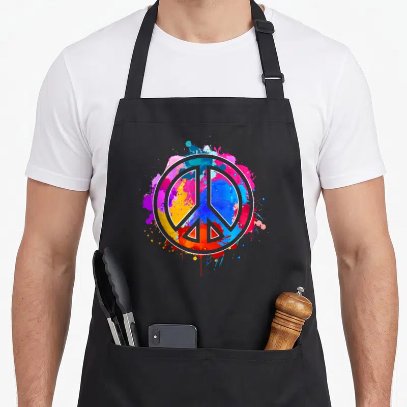 Tablier de cuisine noir avec un grand signe de paix aux éclats de peinture tie dye multicolores sur fond gris.
