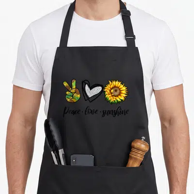 Tablier de cuisine noir avec signe de la paix tournesol et inscription Peace love sunshine en calligraphie.